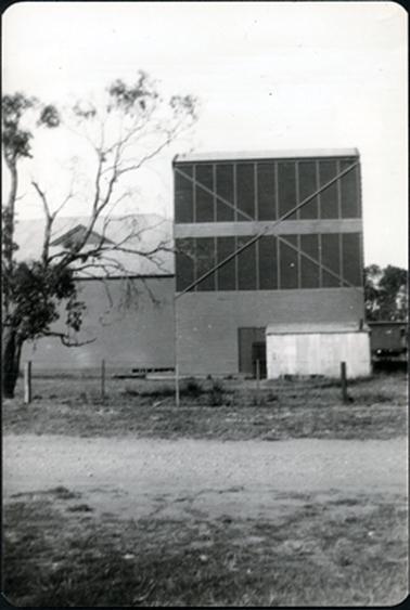 This image shows a large factory-like building behind a post and wire fence.  A gravel road is in the foreground and a tree is on the left-hand side of the image.  A small light coloured building is in front of the factory.  The high section of the factory has several vertical strips and a cross section on the front.  Part of a building with tall trees behind it can be seen on the right-hand side of the image.  