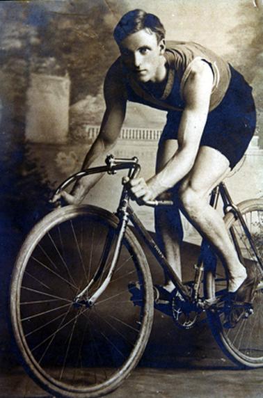 This image shows a studio photo of a young man sitting on a bicycle.  He is wearing black bicycle shorts, a sleeveless top and bicycle shoes which are strapped to the pedals. His hair is parted on the left.  He is leaning forward and gripping the handlebars which are turned upside down.  He is looking at the camera with a serious expression on his face.  The studio backdrop is behind him.  