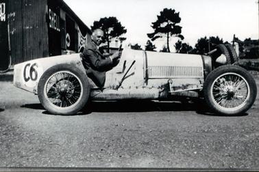 This image shows a man sitting in a racing car and smiling for the camera.  The hair on the sides of his head is grey and he is wearing a white shirt and a suit coat.  The car has ‘C6’ painted on the side at the rear.  Two spare tyres are mounted at the front of the vehicle.  A long lever is on the side of the vehicle near the running board.  Part of a galvanized iron shed is behind the racing car on the left-hand side of the image.  The words ‘shell’ and ‘castrol’ can be seen on the side of the shed.  Tall trees are in the background and gravel on the ground in the foreground.  