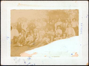 This image shows a large group of twenty-seven men and boys.  There are eleven men in the back row, nine men in the middle row and seven men and boys sitting on the grass in the front row.  Most of them are holding spades.  The image is very faded and a large piece has been torn away from the bottom and right-hand side of the photograph.  There are trees in the background.  There is an embossed border around the mounting board.  