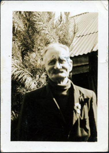 This image shows an elderly man, from the waist up, posing for a photo.  He has white hair and a white moustache.  He is wearing a roll-neck sweater under a coat.  A badge is pinned to his left lapel.  The iron roof of a building is behind him on the right-hand side and a tree is on the left-hand side. 