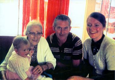 This image shows a group of three adults and a baby posing for a photograph.  The baby is sitting on the elderly woman’s lap. The baby is wearing a white knitted cardigan and a pink dress.  The elderly woman is wearing a white cardigan over a dark printed dress.  She has grey hair pulled back and is wearing spectacles.  The man is sitting between the two women and he is wearing a stripped shirt.  The younger woman on the right is wearing a short-sleeved light coloured cardigan, buttoned up, over a dark dress.  Her hair is pulled back. Windows with orange curtains are in the background.  