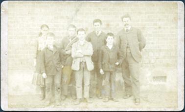 This image shows a man and six children posing for a photograph in front of a brick wall.  A vent can be seen on the right-hand side of the building, near ground level.  The man is dressed in a three-piece suit and has a moustache.  There is a girl and two boys in the back row and three boys in the front row.  They are all wearing coats and boots.  
