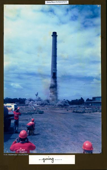 This image shows a tall chimney stack with a door in the centre and a ladder along the right-hand side from the bottom to the top.  Grey smoke swirls around the base.  Four men wearing red helmets and red long-sleeved shirts with the words ‘Able Blasting’ printed on the back are watching the blast.  One man is holding onto a German Shepherd dog.  A silver tank on a small tower is to the left of the chimney stack.  Part of a white van can be seen on the left-hand side and part of two storey building brick building with windows across the front can be seen on the far right-hand side of the image.  Trees are in the background.  Written in black ink on white paper at the bottom of the mounting board is: “..going....”. ‘D.M. Thompson Collection’ is printed in silver at the bottom.