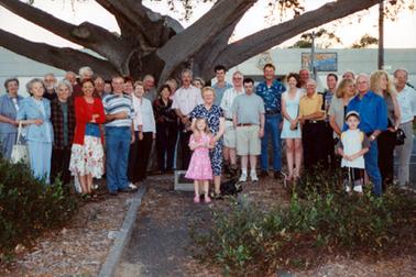 This image shows a large group of people standing under a very large tree, posing for a photo.  Part of six very large branches can be seen extending out from the trunk of the tree.  There are thirty-one adults and two children.  A Marker is on the ground behind the little girl.  A concrete path and small shrubs are in the foreground and part of an office building can be seen in the background.  
