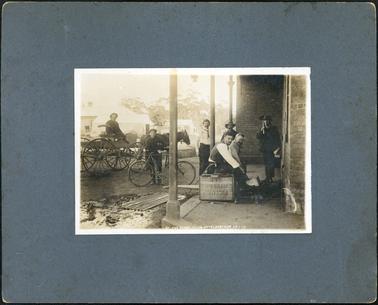 This image shows a group of seven men outside a building.  The building is brick and there are three verandah posts visible.  One man is sitting on a box which bears the inscription “Victoria Bitter Ale”.  One man is standing beside a bicycle.  He has a pipe in his mouth.  Behind him is a man sitting in a four-wheeled wagon with a horse.  Three other men are standing and another is sitting.  A piece of galvanised iron is lying on the ground and there are buildings in the background.  Printed in white on the bottom of the photograph is: “At the Pumps Club Hotel Hastings 25-7-13”.