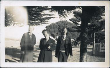 This image shows two women and a man standing on a roadway beside the water.  The taller woman on the left is wearing a cardigan with a belt and a box pleated skirt.  The woman in the centre is wearing an overcoat and holding a bag under her arm.  The man is wearing a shirt, tie and three-piece suit.  There are trees beside and behind them and a jetty can be seen disappearing out into the water.  Part of a building can be seen under the trees in the distance.  