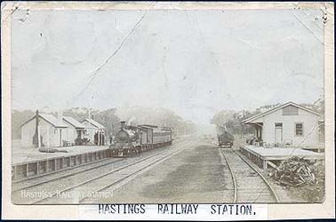 Image of the Hastings Railway Station showing three small wooden buildings on the left platform and one wooden building on the right platform. There are three railway tracks with a steam engine and carriages on the left platform with a group of people standing near the train. There is a wagon on the right hand track. There is a wood pile beside the right hand platform.