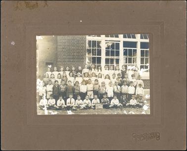 This image shows four rows of school children posing for a photograph outside their school.  There are twelve children sitting on the ground in the front row, and fifteen children standing in each of the other three rows.  The small board on the ground in front of the children reads: “Hastings School No. 1098 Grades I, II, III, IV.”  The school wall behind the children is brick and four very large windows are seen.  The photographers details are embossed on the mounting.   