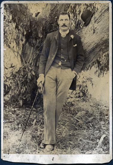 This full-length image shows a well-dressed man leaning nonchalantly against the trunk of a large gum tree.  He is wearing a three-piece suit.  His shirt has a small collar and he is wearing a cravat.  He has a flower in his coat lapel, a handkerchief in his coat pocket and a fob watch in his waistcoat.  He is holding a walking stick and gloves in his left hand and his right hand is in his pinstriped trouser pocket.  His legs are crossed and a top hat is resting on the tree trunk.  He is standing on overgrown grass and leaves.  