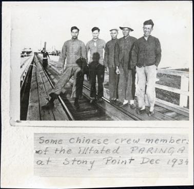 This image shows five Chinese men standing on a railway line on a jetty, posing for a photograph.  Another man is standing on the railway line in the background.  The man on the left-hand side of the photograph has his right leg out to the side.  All are wearing long sleeved jackets and dark trousers, except the man on the right, who is wearing light coloured trousers.  One man is wearing a hat and one a cap.  There are wooden rails on either side of the jetty.  Boats can be seen in the far background.  Printed at the bottom of the image is: “Some Chinese crew members of the illfated PARINGA at Stony Point Dec 1934.