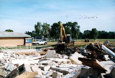 This image shows a demolition site.  A large number of broken-up concrete blocks are on the ground.  A man working an excavator can be seen in the centre of the image.  A brick building with a galvanised iron roof and a painted mural, is on the left-hand side.  A white utility is parked beside it and children’s playground equipment can be seen behind it.  A machinery scoop is sitting on the grass to the right of the excavator.  White pipe railings are on the left-hand side of the image.  Tall trees, grass and part of another building are behind a tall wire fence in the background.  