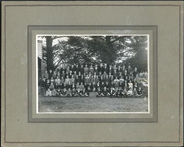 This image shows a group of school children, in five rows, posing for a photograph, under large pine trees.  Part of a timber building can be seen on the far left-hand side of the photograph.  There are seventeen children in the back row, nineteen children in the second back row, twenty children in the middle row, twenty-one children in the second front row and nineteen children sitting, cross-legged on the ground, in the front row.  A boy in the front row is holding a small blackboard which reads: “Hastings School 1098 -.1935.-”.  