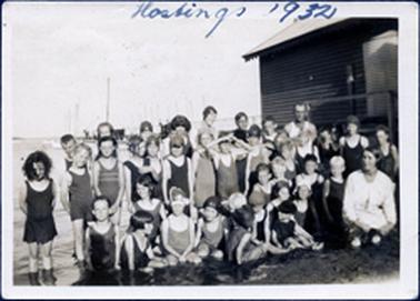 This image shows a large group of approximately forty children and three adults posing for a photograph, in water, beside a shed.  All the children are in swimming attire and either sitting or standing.  A woman, dressed in white, is squatting in the front row on the far right-hand side.  Another woman and a man are standing in the back row.  A jetty is in the background.  Boats and tall masts can be seen beside the jetty.  Written at the top of the image in pen is: “Hastings 1932”.  