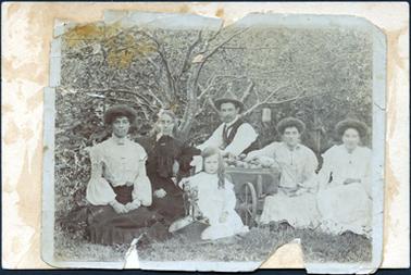 This image shows a family group sitting on the grass in front of fruit trees, posing for a photograph.  There are two women on the left, one young and the other older.  The young woman is wearing a white long sleeved blouse with high lace neck and large puffed sleeves from the elbow to the wrist.  She is wearing a dark coloured skirt.  The older woman is wearing dark coloured clothes with a large frill at the front of her bodice.  The two young woman on the right of the image are wearing white clothes, both with long sleeves and high neck lines.  All women are wearing brooches at the base of their necks.  There is a man in the centre of the image leaning on a wheelbarrow full of apples.  He is wearing a high collared, long sleeved white shirt, a waistcoat and a hat.  A small girl is sitting at the front holding the handle of a basket full of apples.  She is dressed all in white and has curls over her left shoulder and a large white bow on the opposite side of her head.  Everyone is beautifully dressed. There is grass in the foreground.  Fruit can be seen on the trees in the background.  The photograph has been glued onto a piece of card.  