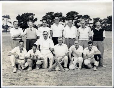 This image shows a group of thirteen men, on grass, posing for a photograph.  There are seven men standing in the back row and six men crouching or kneeling in the front row.  All the men in the front row are wearing ‘whites’.  The two men in the centre are holding a cricket bat each, crossed in front of them.  The man second from the left is wearing cricket pads and gloves.  The men in the back row are wearing white shirts and different coloured trousers.  Two of these men are wearing sleeveless pullovers.  The man wearing the tie has only one arm.  Tall pine trees are in the background.  
