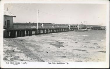 This image shows a pier jutting out into the water with a sandy foreshore in the foreground.  Boats are on either side of the pier and several people are walking along it.  Power lines are running alongside the pier between four tall white poles.  Part of a shed is visible on the far left-hand side of the image.  Land and trees can be seen in the distance.  Printed across the bottom is: “The Rose Series P.10375 Copyright The Pier, Hastings, Vic.”