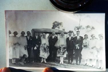 This image shows a wedding party consisting of eight women and five men.  The two brides appear to be dressed identically in knee length dresses, long sleeves, high neck with small collars and elbow length veils.  They are both carrying very large bouquets and a horseshoe.  An elderly man is standing between the brides.  Two men are on either side of the brides and three bridesmaids are on either end of the group.  The men are all wearing three piece suits and the bridesmaids are wearing knee length dresses with three quarter length sleeves.  They are wearing black shoes with a strap across the top.  A motor vehicle is behind the group and a dog is sitting at the front, on the left-hand side.  Two trees can be seen in the background.  