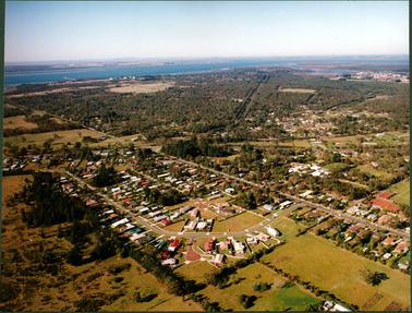 This image shows an aerial photograph of a small town.  Buildings and streets are in the foreground and the foreshore and sea are in the background.  Land can be seen in the far distance.  A large section of land at the centre of the image is heavily treed.  A jetty and tanks can be seen in the distance on the left-hand side.  A long straight road runs across the centre of the image.   