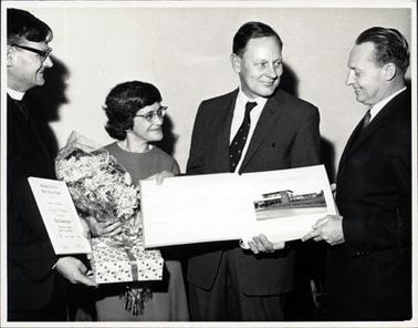 This image shows a group of three men and one woman.  The three people on the left are looking at the man on the right who is presenting the man next to him with a book.  Both these men are dressed in suits, white shirts and ties.  The man on the left-hand side of the image is a minister of religion and he is holding a certificate.  The woman is holding a present in a box tied with ribbon and a bouquet of flowers.  Everyone is smiling.  