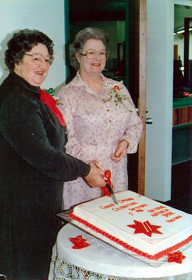 This image shows two women standing in front of a large cake.  They are both holding a knife, tied with a red ribbon and are ready to ‘cut the cake’. The cake is iced in white with red writing and decoration.  The writing reads: ‘Grand Opening Celebrations September 1st 1986  National Australia Bank Hastings’. The cake is sitting on a white lace tablecloth on a round table.  Both ladies are wearing spectacles and have their hair neatly set.  The lady on the left is wearing a dark green coat with a red tie at the neck.  The lady on the right is wearing a long sleeved print dress.  Both ladies have a corsage pinned on the left-hand side of their outfits.  They are both smiling.  An open doorway is behind them.  