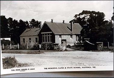 This image is of the Memorial Gates and the State School at Hastings.  It shows the main building with four large windows at the front and a chimney on the right-hand side.  An older building is on the left-hand side.  The Memorial Gates are curved stone entry with two wire gates.  There is a letterbox on the footpath and the road is gravel.  Many large trees are beside and behind the buildings.   The title is printed at the bottom of the photograph along with the maker’s details.  