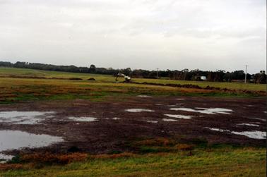 This image shows a large grassed paddock with puddles of water lying in the foreground.  Tall trees are in the background.  An excavator is digging a trench across the paddock at the centre of the image.  Part of a building and two electricity poles can be seen in the background on the right-hand side.  