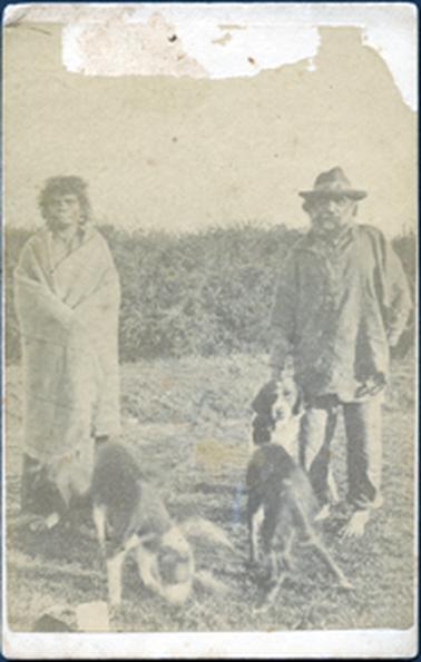 This image shows an aboriginal woman and man standing on grass looking at the camera.  The woman has a blanket wrapped around her shoulders and appears to be barefoot.  She has curly hair to her shoulders.  The man is wearing a long sleeved shirt hanging out over long pants which are rolled up at the bottom.  He is wearing a hat and is barefoot.  He has a beard and moustache.  There are two dogs in the foreground and one dog standing beside the man.  There are thick shrubs behind them. 