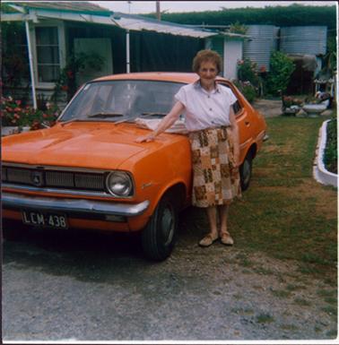 This image shows a woman standing beside an orange car, posing for a photograph.  She is wearing a short sleeved white blouse, a patterned skirt and lace-up shoes.  She has short, curly hair and is wearing spectacles.  One hand is resting on the car which has the licence plate number LCM438.  The car is parked on the lawn in front of a house.  Gardens can be seen on either side of the photograph and two large galvanised iron water tanks are in the background.  