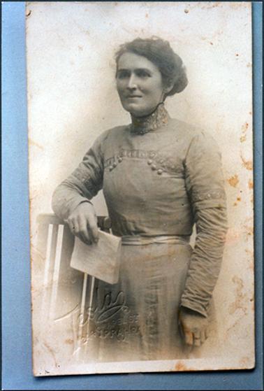 This image shows a woman standing with her right arm resting on the back of a wooden chair.  She is wearing a long sleeved dress with a waistband with a very high embroided neckline. There are six buttons  on the bodice of her dress and there is a small brooch at the neck.  She is wearing a ring on the middle finger of her left hand and her hair is swept up at the back.  She is holding a piece of paper in her right hand.  The photographers details are on the left-hand side.