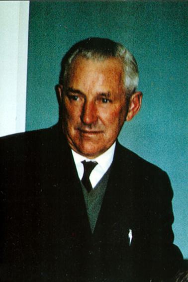 This is a photo of a man, taken from the waist up.  His neat, greying hair is parted on the left side.  He is wearing a white shirt, black tie, green vest and black coat.  He is looking away from the camera.  A blank wall is behind him.  