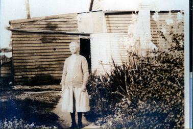 This image shows an elderly woman standing on a path in front of a wooden building.  She has grey hair and is wearing a long sleeved cardigan over a knee length dress.  She is wearing black stocking and shoes.  She is looking slightly to the left and her arms are by her side.  A galvanized iron rain water tank is in front of the building. Shrubs are on the right-hand side of the path.  