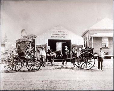 This image shows a corrugated iron workshop with the words “W. SMITH GENERAL BLACKSMITH WHEELWRIGHT & [...]” To the right of the workshop a timber house with a verandah can partially be seen. In front of the workshop are a covered buggy with 4 wheels and a 2 wheeled cart with the words “F.G. PERROTT BUTCHER” written on the side. 4 men are standing around the cart, one with a bundle tied to a stick over his shoulder, and a small girl on a pony.