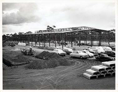 This image shows men working on a large steel framework for a building.  Cars are parked in the foreground.  There is a Volkswagon, 2 Ford Falcons, a Vauxhall, Plymouth, Buick and an MG.
