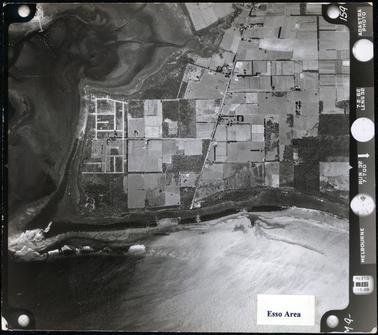 This image shows an aerial view of land jutting out to a point, with water on two sides.  The land is broken up into many small paddocks and some houses can be seen dotted around.  A bay on the far left-hand side of the image shows a jetty and boats, which look like little white specks, dotted on the water.  A piece of white paper with “Esso Area” typed in black, is glued to the bottom of the image.  Photographic information in white print, is down the right-hand side of the image.  