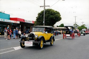 This image shows four vintage motor vehicles, followed by fire trucks, driving down the middle of a street.  People and shops line the left-hand side of the street.  Two of the shops are “Home Hardware” and “ACLSORTS”. The first vehicle is yellow with black mud guards, four doors, a black soft top and yellow spokes in the wheels.  Three people can be seen inside the vehicle. The second vehicle is black with a black soft top and very large red wheels.  Two people are in the vehicle.  A small Australian flag is on the vehicle.  The next vehicle is black with large red wheels but has no top.  It carries two people and the last vintage motor vehicle is green with a black soft top.  Fire trucks follow.  All cars have a sign at the front which reads: “Hastings-Western Port Historical Society Incorporated”.  Several power poles line the street and a large tree is behind the yellow car.  