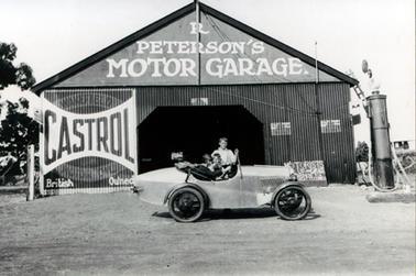 This image shows a small racing car parked in front of a garage.  A woman is behind the wheel and a small boy is sitting next to her.  Another boy is standing behind them.  The car is parked in front of a large galvanized iron shed with the following signs painted on it: ‘R Peterson’s Motor Garage. Castrol Wakefield Motor Oil British Owned Motorists up and garage here via boat to Cowes’.  A petrol bowser is in front of the  garage on the far right-hand side and a ladder can be seen behind the bowser leaning against the garage.  Branches of a large tree can be seen on the far left-hand side.  