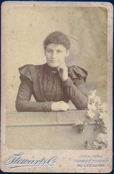 This image shows a young woman, from the waist up, posing for a photograph.  She is standing behind a waist-high wall, with both arms resting on top of the wall.  Her left arm is bent and her hand is resting on the left side of her neck.  He hair is pulled back.  She is wearing a dark coloured dress.  It has a self embroided pattern,  long sleeves with a turned back cuff.  There is an additional small pleated capped sleeve.  There are pin-tucks across the front of the dress and along the large, deep lapels on the dress.  A bar brooch is at the base of her neck.  She has a very slight smile on her face.  Flowers can be seen in the foreground on the right-hand side.  The photographers particulars are embossed in silver at the bottom of the mounting board.  
