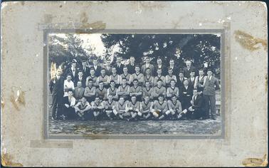This image shows the Somerville Premiership Football Team of 1929 posing for a photo.  There are forty-five men standing and sitting in four rows.  All the players have their arms folded and are wearing light coloured, V-neck T-shirts with a dark colour V at the neck, around their shirt cuffs and at the top of their socks.   A football with “Premiers 1928 M.P.F.A.” is on the ground at the front between the players.  All the other men are dressed in three piece suits, white shirts and ties.  Tall trees are in the background.  
