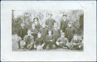This image shows a group of thirteen young men and boys, in three rows, posing for a photograph. Three are standing at the back, five kneeling in the middle and five sitting on the grass.  Two men in the back row are wearing hats and the third is wearing a cap.  Four others are holding a hat in their hands.  All are well dressed, some in three piece suits.  Two boys on the right-hand side in the front row have short pants and long socks.  There are trees in the background and part of a dog can be seen on the far right side of the image.  