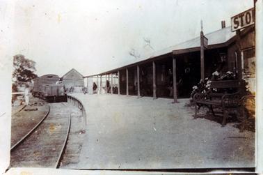 This image shows a railway station that has a verandah with a row of posts across the front.  It has a sloping galvanized iron roof.  Several people can be seen sitting and standing under the verandah.  A sign post is on the right-hand side of the image behind a flowering shrub and a bench seat.  Part of a large sign with the letters ‘STO’ can be seen on the far right-hand side.  A railway line and the back of a goods train is on the left-hand side of the image.  Part of another building can be seen between the train and the station.  A large tree is at the left of the train.  