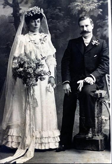 This image is a studio portrait of a bride and groom.  The bride is standing.  Her dress is to the floor and has long sleeves puffed above the elbow and straight to the wrist.  A small band and lace frill is below the elbow.  There are three rows of frills around the bottom of the skirt.  The neckline is high, with a brooch and there is an overlay of lace on the bodice.  There is a corsage on the left-hand side of her bodice.  She is carrying a large bouquet of flowers tied with a very large ribbon which is falling across the floor.  She is carrying gloves in her left hand.  Her headpiece is made up of tiny ‘teardrops’ and her veil falls almost to the floor.  The groom is half-sitting on a pedestal with one foot on the floor.  He is wearing a three-piece suit with a very high collar and a small bow tie.  He has a spray of flowers in his coat lapel.  He is holding gloves in his left hand.  He has a large moustache.  The wall behind has images of trees and leaves.  
