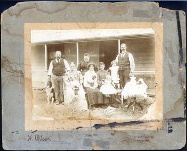 This image is a portrait of a group of two men, four women, one child, one baby and a dog.  Both men have long-sleeved shirts, ties and vests.  The baby is sitting on the lap of the woman to right of the photo and the child is standing on a frame beside the man on the right-hand side of the photo.  The dog is a border collie.  There is a small table with a cloth and refreshments on the right-hand side of the photograph and a teapot, with a tea cosy,  is on the ground.  The group is standing in front of a weather-board house with a galvanized iron roof and verandah posts.  There is a mesh fence around the verandah. Printed on the bottom of the image is: “H. Wilson, 54 Greville St., Prahran.”. 