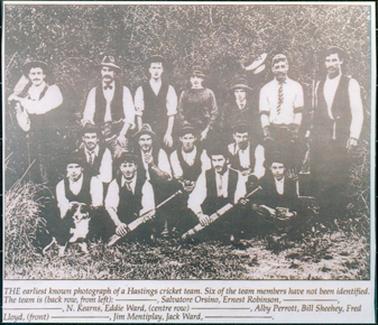 This image shows a group of fifteen cricketers, in three rows, posing for a photo. There are seven men standing in the back row, four men crouching in the middle row and four men sitting on the grass in the front row.   All the men are wearing hats or caps.  Most have white, long sleeved shirts and ties.  A man on the left-hand side in the back row is holding a cricket bat over his shoulder.  The man, second from the right in the back row, is wearing cricket gloves and leg pads.  Two men in the front row are holding cricket bats.  A border collie dog is sitting at the front of the men.  There are large shrubs behind the men.  The names of the men are printed under the photo.  
