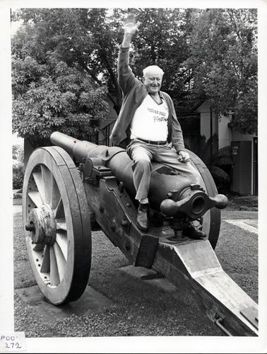 This image shows a very happy grey haired man sitting on the top of a gun barrel waving his right hand for the camera.  He is holding a rag in his left hand.  He is dressed in light coloured trousers, a white T-shirt with the words ‘Western Port Festival’ on the front and a open cardigan. The gun is sitting on a field carriage on the lawn in front of a building.  There is a large tree in front of the building and a tree fern on the right-hand side in the background.  