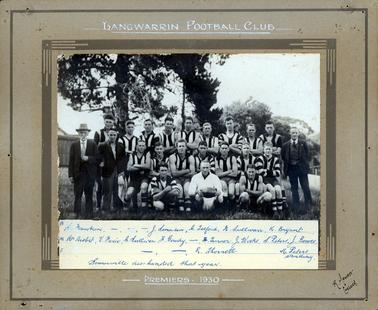 This image is of the Langwarrin Football Club posing for a photograph in a grassed paddock in front of a large pine tree.  There are trees in the background and parts of fences and buildings can be seen on either side of the image.  There are nine men standing in the back row, seven men sitting in the middle row and three men squatting in the front row.  There are two men standing on the left-hand side of the image, one elderly and one young.  They are both wearing suits.  One man, wearing a three-piece suit is standing on the right-hand side of the image.  The man squatting in the centre, front, is dressed in white and holding a football.  Printed in white on the mounting at the top is:” Langwarrin Football Club”.  Printed in white at the bottom of the mounting is: “Premiers. 1930”.  Written on paper in blue biro and glued to the bottom of the image is: “(B)  E. Hawken, ___, ___, ___, J. Swanson, A. Telford, H. Sullivan, K. Bryant.(M) Mr Hisbit, V. Fasio, A. Sullivan, F. Goudy, ___, H. Turner, J. Wicks, S. Peters, J. Bowes, (F) ___, ___, R. Thornell, M. Peters, standing.” Also written below the names is: “Somerville disbanded that year.”. The photographers name and address is printed on the bottom right-hand corner of the mounting.  