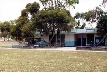 This image shows part of a long low building with a flat roof.  There are full length windows at the centre of the building.  Smaller sets of windows are on the far left-hand side and doors on the far right-hand side of the building.  The bottom section of the walls on either side of the full-length windows has a wavy blue pattern.  Trees are in front of and behind the building.  Wooden seating is in front of the building.  Wooden rails are around the trees.  A fence and a white car can be seen in the distance on the far left-hand side.   