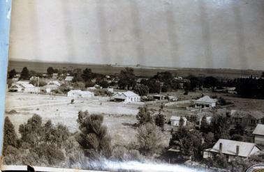 This image shows a township taken from an elevated position.  There is undeveloped land on the left and houses on the right and across the middle of the image, set amongst the trees.  There is a large double gabled building in the centre with a street running in front of it. Trees are in the foreground and water can be seen in the background.  Land can be seen in the far background.  