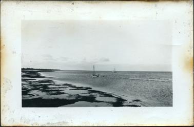 This image shows a foreshore at low tide.  Two small boats with masts can be seen in the centre of the image.  Two small waves can be seen on the right-hand side and sand and seaweed is on the left-hand side of the image.  Trees can be seen on an outcrop of land in the far distance on the left-hand side of the image.  Low clouds are on the horizon.  The edges of the photograph have brown burn stains.  
