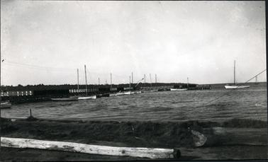 This image shows a photo taken from the foreshore looking across the water where there are several fishing boats tied to a jetty on the left-hand side of the image.  A row of trees can be seen in the distance and a large pole is lying on the ground in the foreground.  